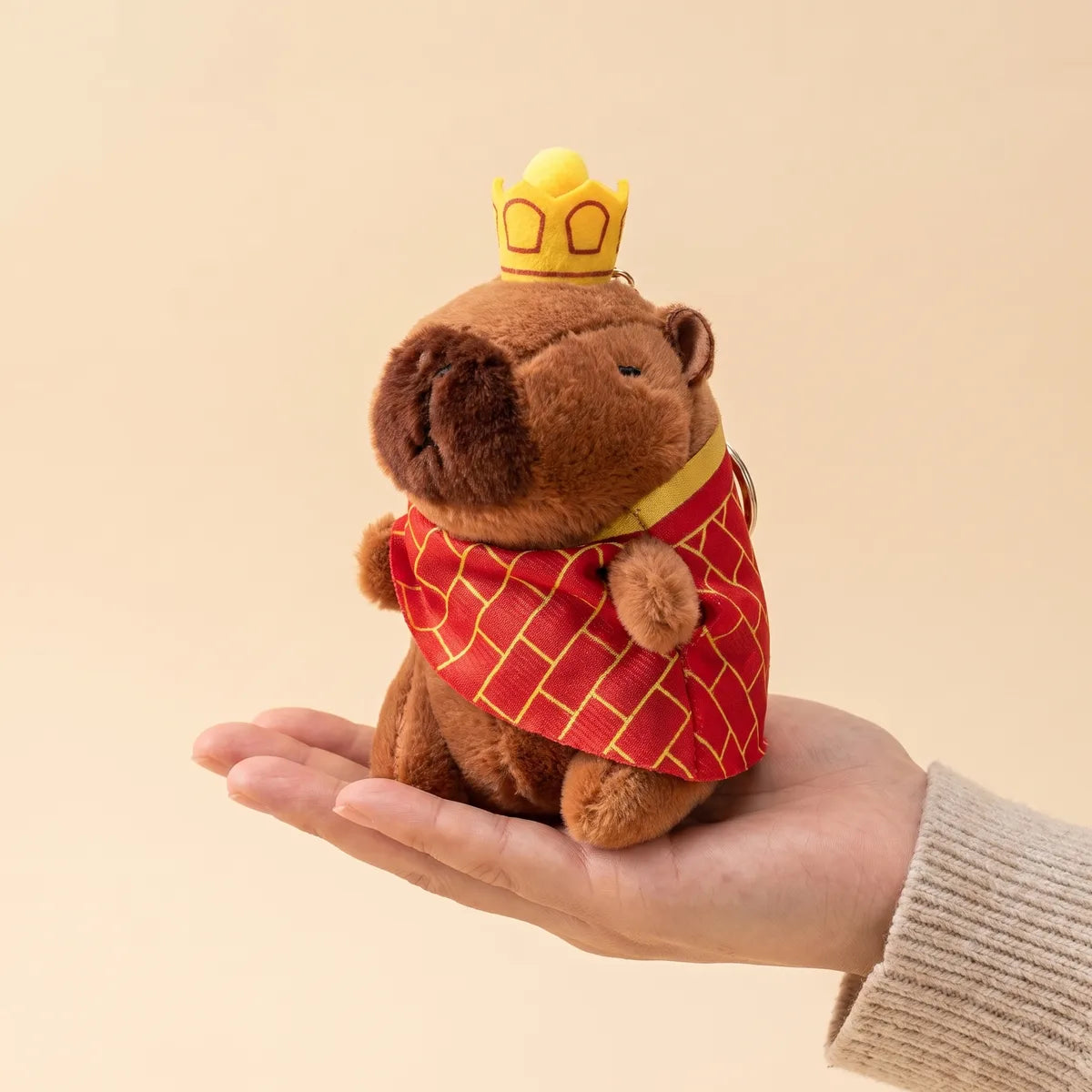 King mini capybara plush standing upright on an open palm with its cape spread over the fingers