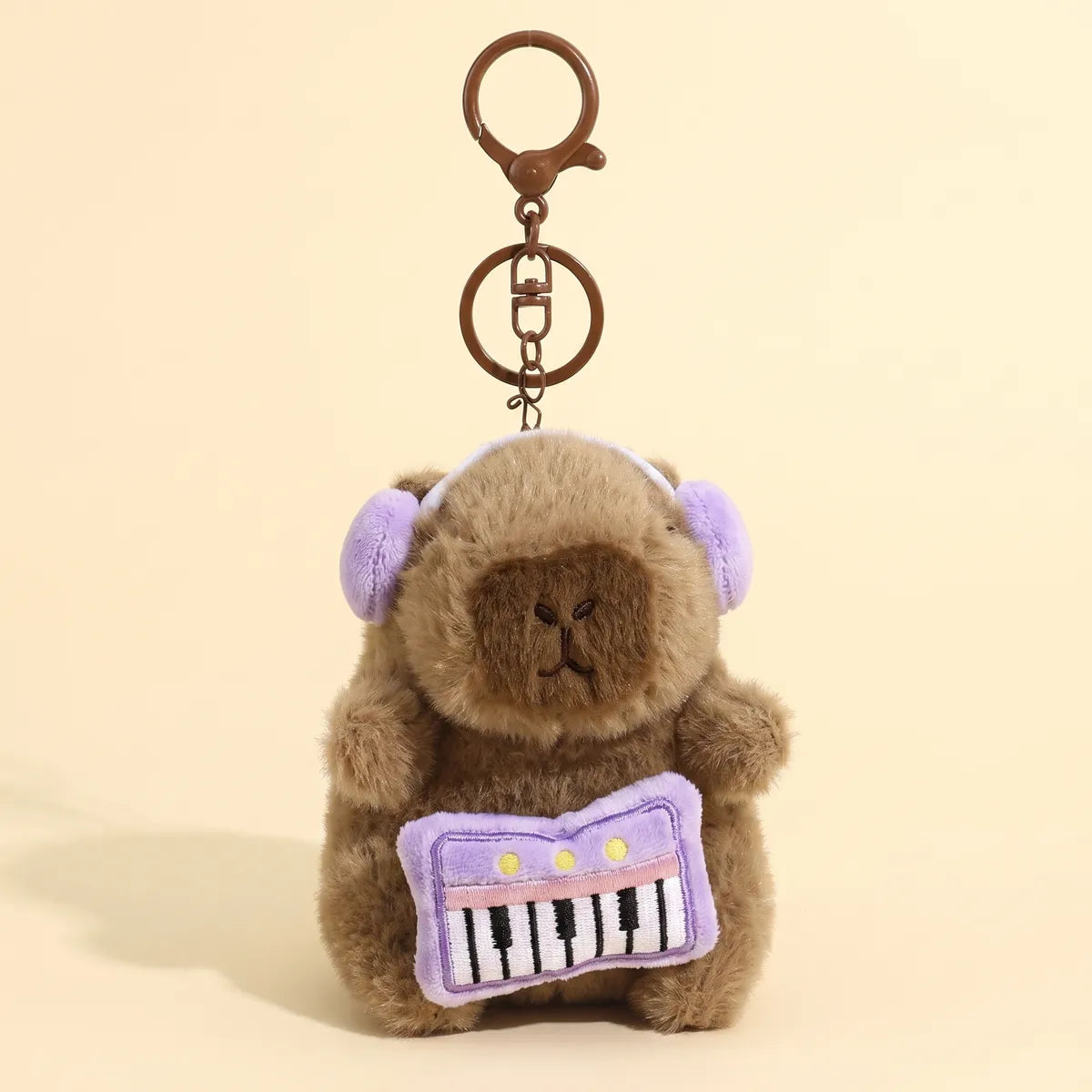 Piano capybara keychain hanging from its bronze clasp with the purple keyboard and lilac headphones facing front