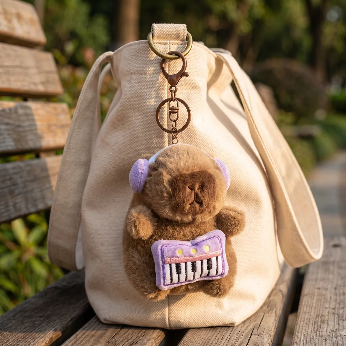 Piano capybara keychain clipped to the handle of a cream canvas tote bag on a park bench