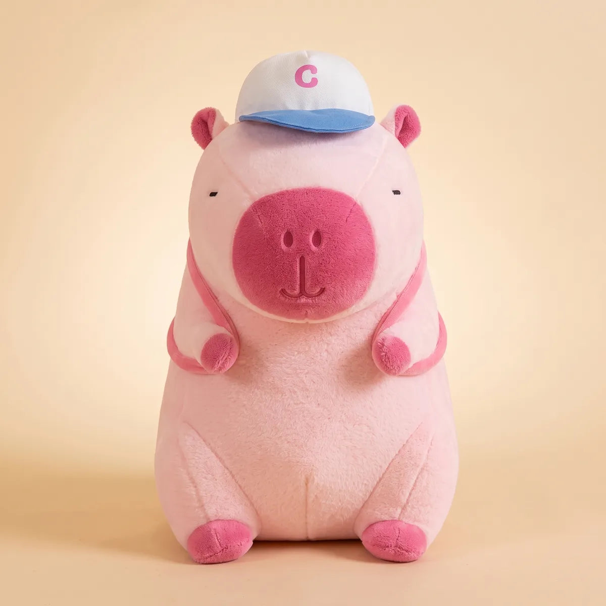 Pink capybara plush seen from the front with its baseball cap and pink backpack straps