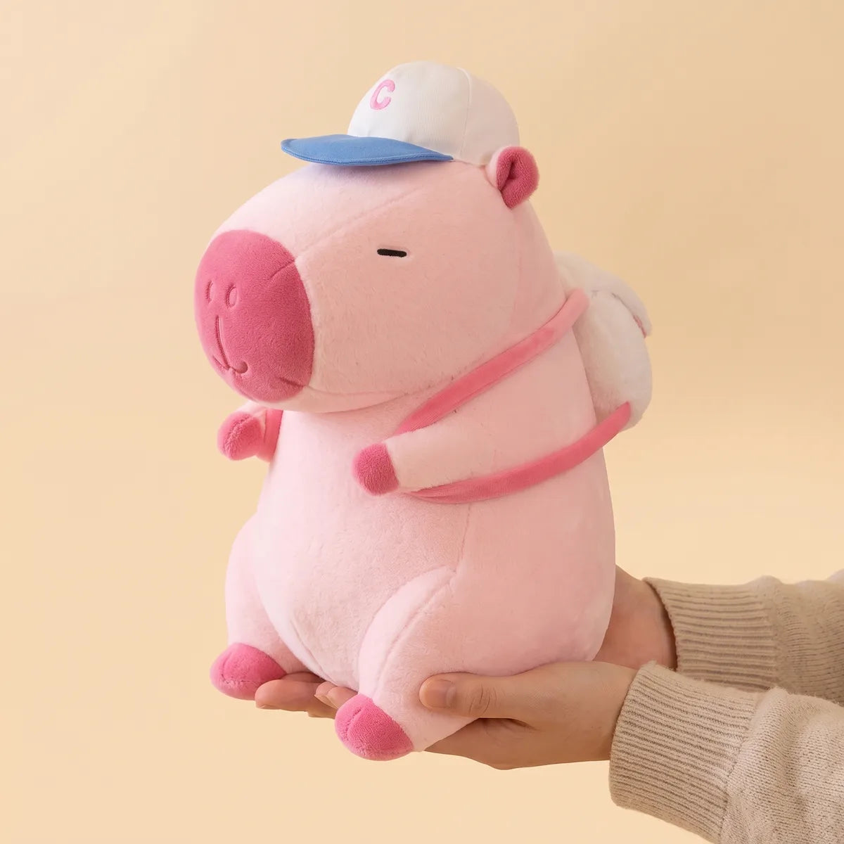 Hands holding the pink baseball backpack capybara plush to show its real size