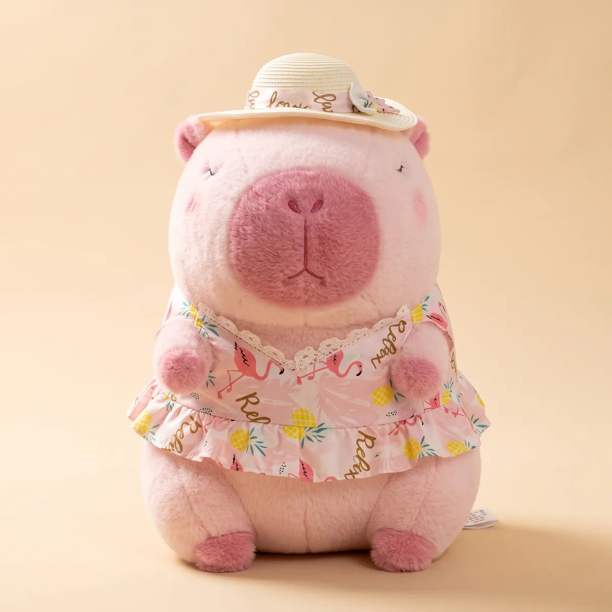 Front view of the seated pink capybara plush wearing its straw sun hat and ruffled flamingo print dress
