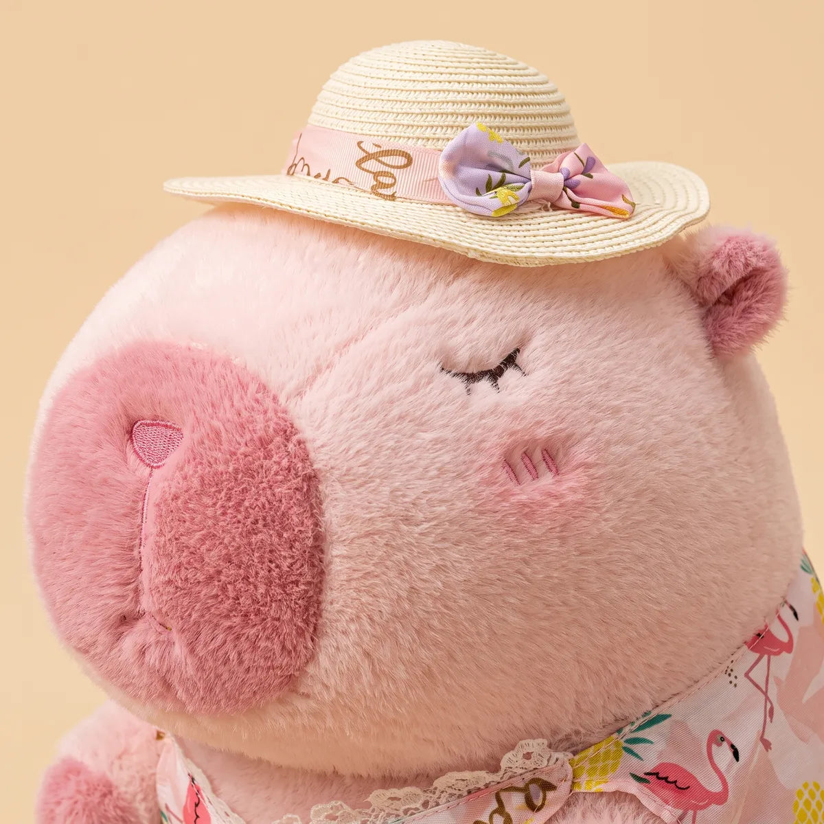 Close-up of the pink capybara plush face with closed stitched eyes, blush cheeks and a lilac bow on the straw hat