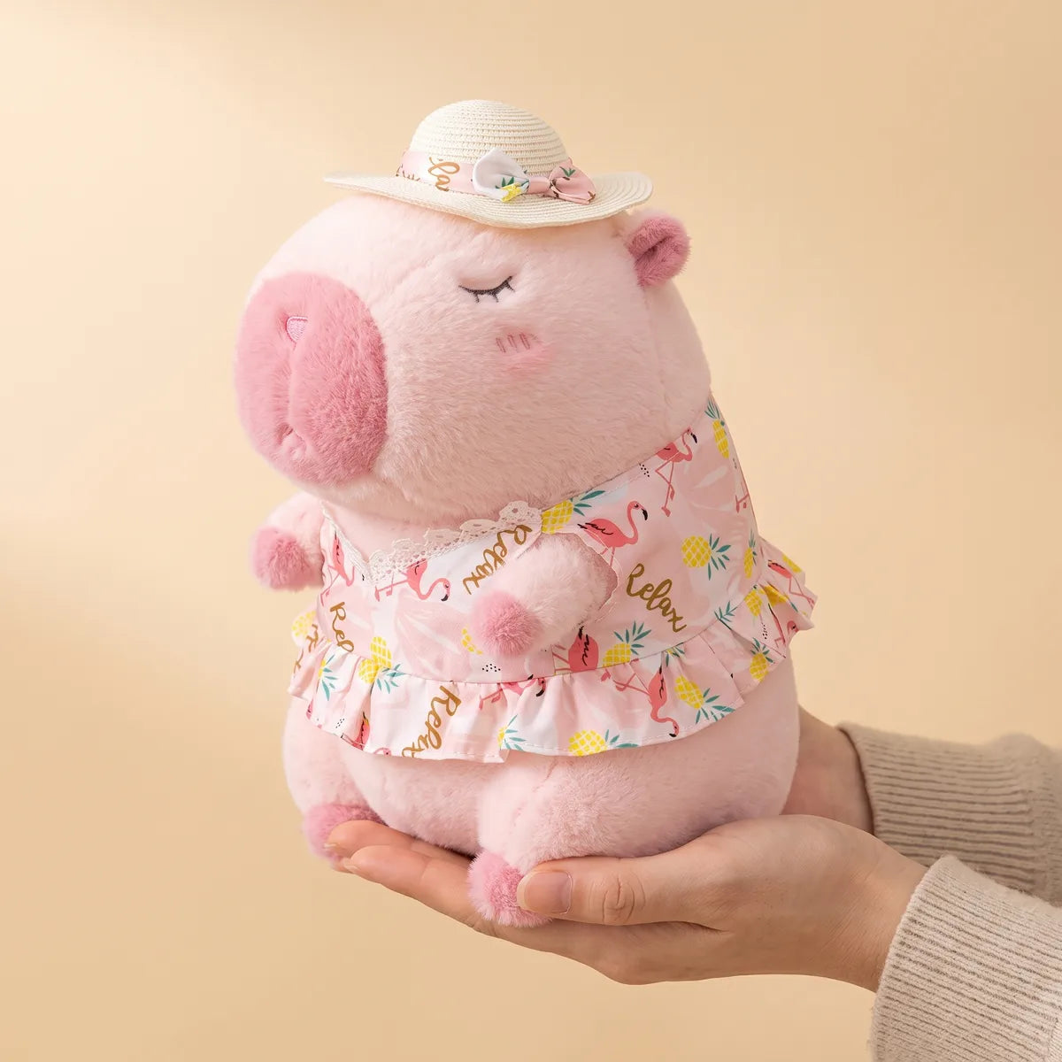 Pink capybara plush in a sun hat resting across two cupped hands to show its 12 in height