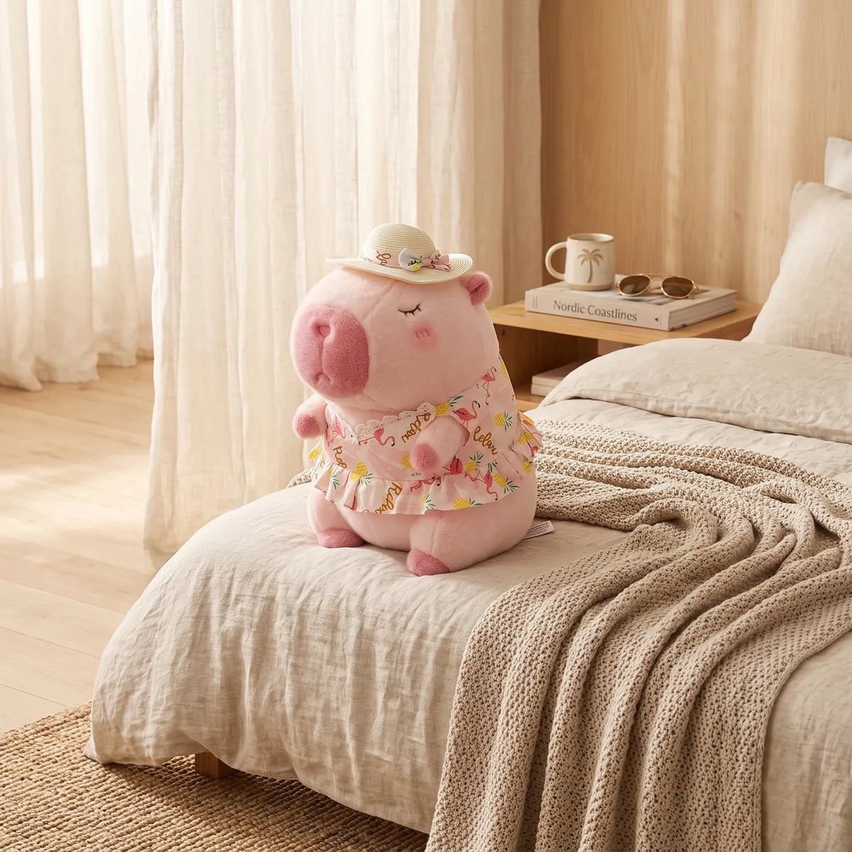 Pink capybara plush in a sun hat perched on the corner of a linen bed beside a nightstand