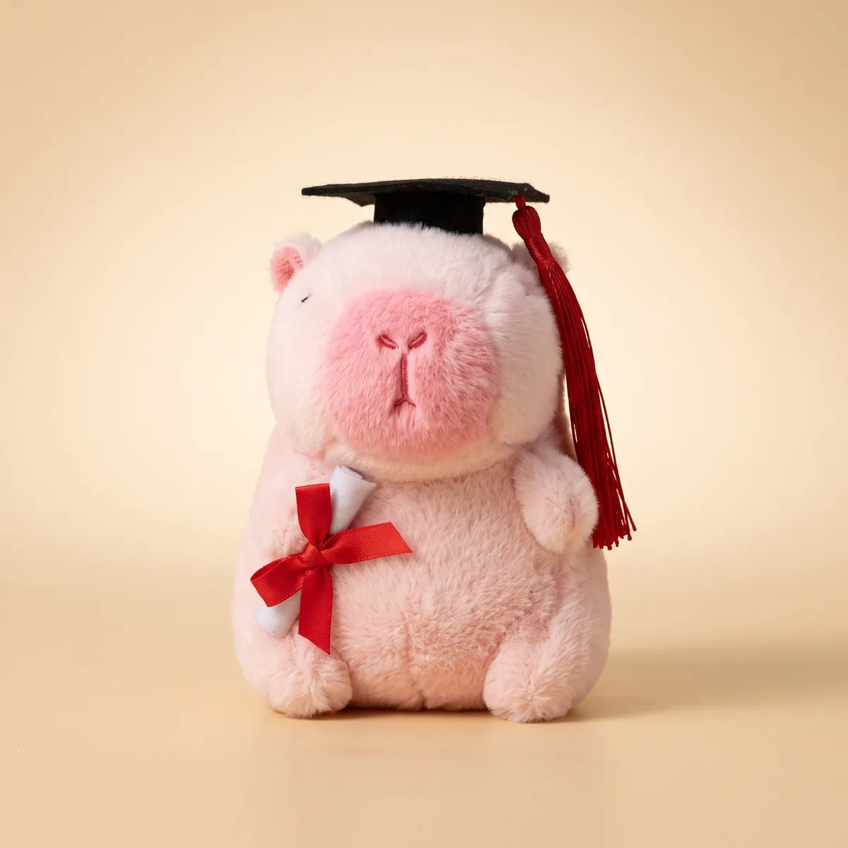 Front view of the pink graduation capybara plush seated on a cream backdrop with its rolled diploma under one arm
