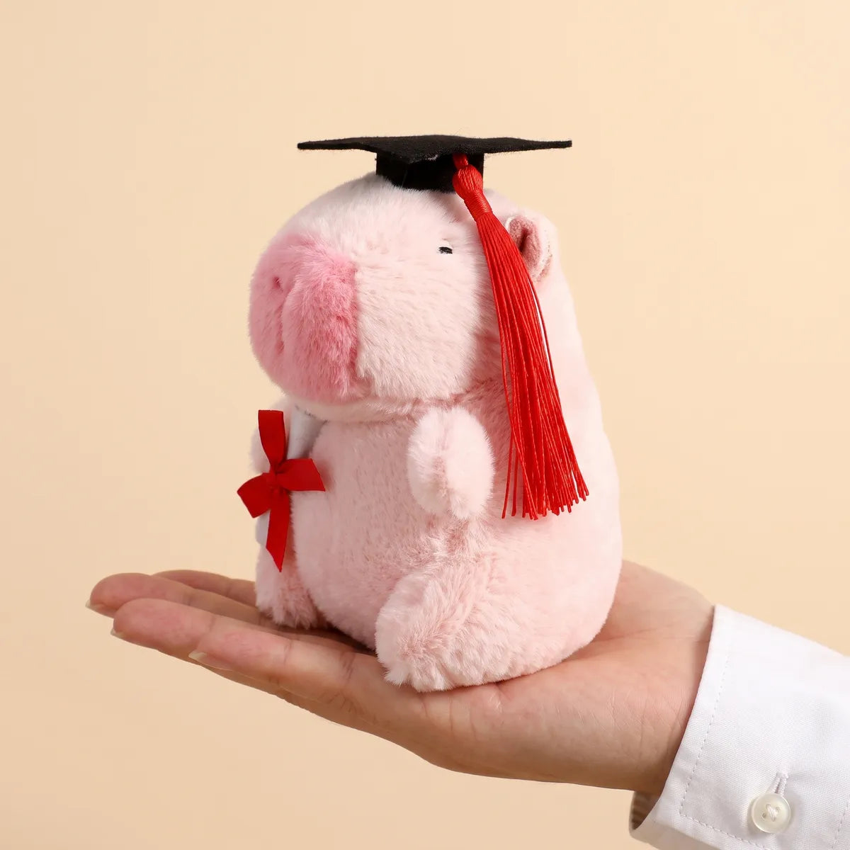 Pink graduation capybara plush resting on an open adult palm to show its 5 in seated height