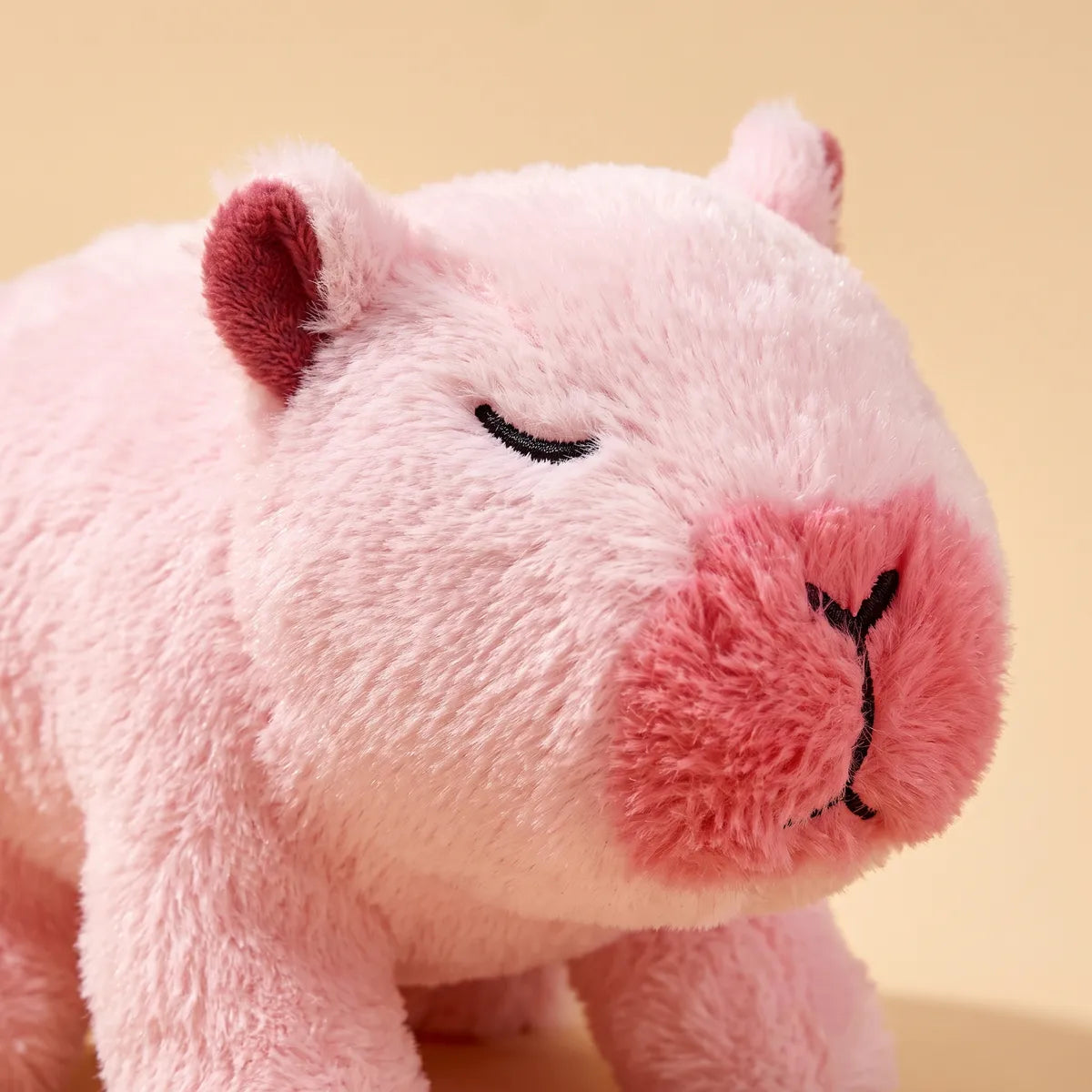 Three quarter head portrait of the pink walking capybara toy, the visible eye stitched closed in black thread above its lashes