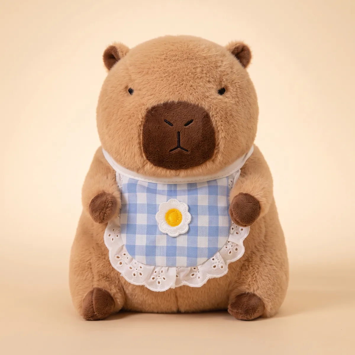 Brown capybara plush seen from the front wearing its blue gingham bib with a daisy