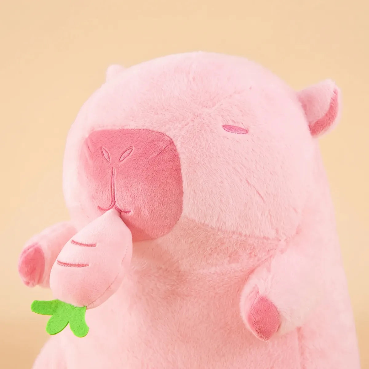 Close-up of the pink capybara plush face with stitched closed eyes and a rose muzzle beside the green turnip top